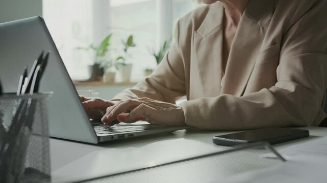 Cropped slowmo shot of female business professional in elegant beige blazer working on laptop at modern desk setup, with office supplies and greenery on windowsill in blurred background