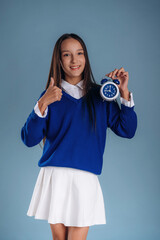 Fototapeta premium With blue clock in hand. Young schoolgirl in uniform is against background