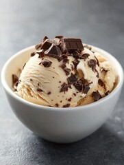 A creamy scoop of ice cream topped with chocolate chunks, served in a white bowl against a dark background.