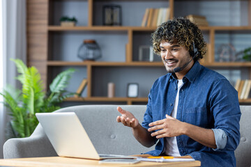 A cheerful man sits on a couch, engaging in a video call. He's wearing a headset, actively...