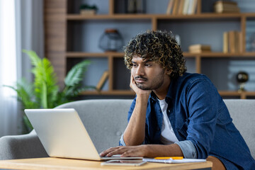 A young man with curly hair appears bored while using a laptop at home. He rests his head in his...