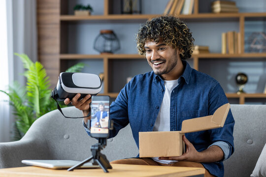 A man enthusiastically unboxes a virtual reality headset during a livestream. He is using a smartphone and tripod to share the experience. The setting is relaxed and modern.