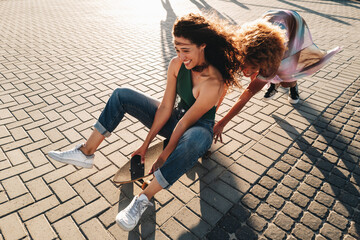 Carefree summer: Playful girl friends pushing each other on a skateboard under the sun