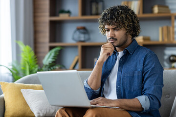 A young man intensely focused on his laptop while seated on a cozy sofa. His thoughtful expression suggests a commitment to work or study. Casual setting with modern decor enhances a relaxed