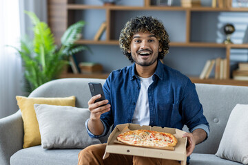 A cheerful man sits on a sofa, holding a smartphone and a pizza box. He appears delighted,...