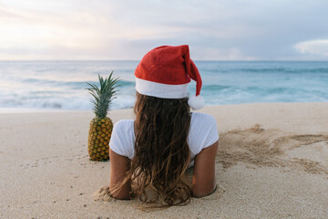 Rear view of a woman wearing a Christmas Santa hat lying on a sandy beach next to a pineapple, Haleiwa, Hawaii, USA