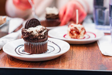 Close-Up of a Delicious Chocolate Cupcake With Oreo Cookie Topping on a White Plate, Perfect for Dessert Lovers and Bakery Enthusiasts, Capturing Indulgence in a Warm and Inviting Café Setting