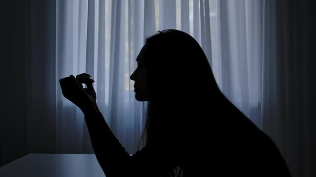 Silhouette of woman sitting at table in front of window, begging regretting expression, sad heartbroken mood, painful tired face. Human emotions and mental health.