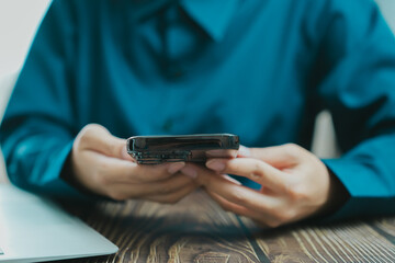 person holding smartphone while sitting at wooden table, wearing blue shirt, with laptop nearby. scene conveys sense of focus and engagement with technology
