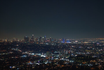 Obraz premium The Los Angeles skyline is seen at night, with skyscrapers brightly lit against a dark sky. City lights form a vibrant tapestry, highlighting the urban landscape.