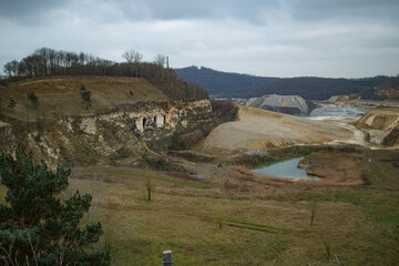 A quarry landscape with exposed earth, piles of excavated material, and a small pond.  Bare trees and  sparse vegetation are visible.