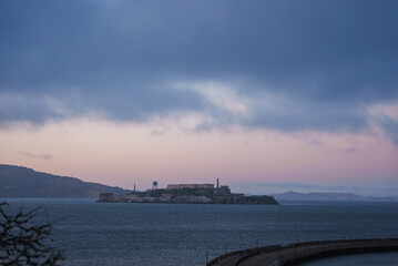 Alcatraz Island is silhouetted against a serene dusk sky with blue and pink hues. Historic prison buildings are visible, surrounded by calm waters and distant hills.