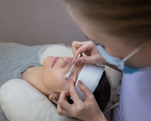 The master applies cleansing foam to the client's eyelashes using a cotton swab. 