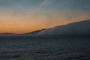 The image shows a tranquil ocean at sunset, with fog enveloping distant mountains. The sky features soft orange and blue hues, typical of the California coast.