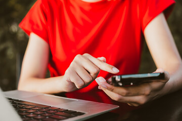 person in red shirt using smartphone while sitting at desk with laptop. scene conveys sense of productivity and modern technology