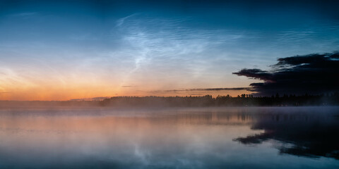Noctilucent clouds illuminating sunrise over misty lake and forest