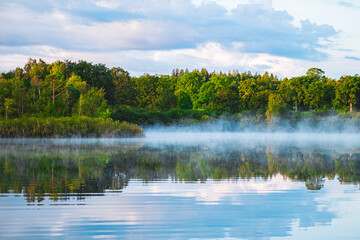 Serene Mist Over Tranquil Lake With Lush Green Forest Reflections Under Beautiful Blue Sky On A Calm Day In Nature