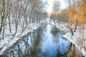 Outstanding landscape in city park with snowy trees and beautiful reflection in frozen river.