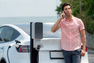 Holiday road trip vacation traveling by the seashore road with electric car, man recharge EV car with green and clean energy while using smartphone. Seascaoe travel with eco-friendly concept.Perpetual
