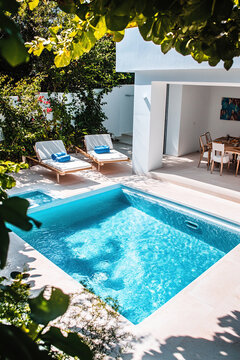 Small square pool in the garden of a Greek villa, surrounded by lush greenery and sun loungers. A dining table is placed on one side with blue pillows. The photo was taken from above.