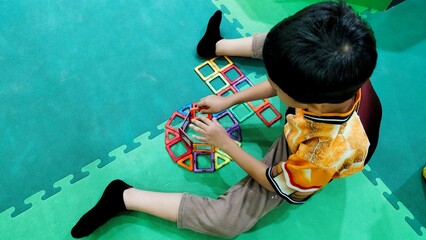 A young boy builds creative structures with vibrant magnetic tiles, seated on a green mat in a playroom