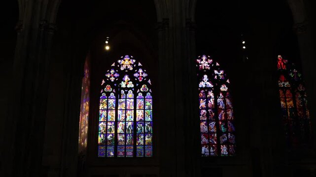 A row of stained glass windows in a cathedral, depicting biblical scenes with vivid colors. The light filtering through creates vibrant reflections, enhancing the solemn atmosphere.