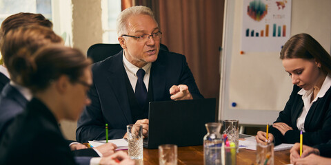 Business elderly leader guides team of professionals through strategy discussion in modern office setting, with charts and graphs displayed. Concept of business, teamwork, leadership, brainstorming