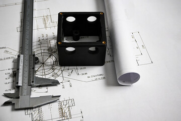 Metal caliper resting on technical engineering drawings with a black plastic part, showcasing precision and manufacturing