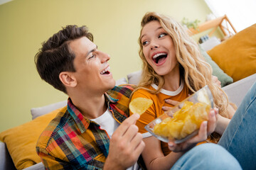 Photo of cute young couple excited eating chips cozy day light spring summer home interior living...