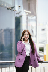 Fototapeta premium A businesswoman stands outdoors amidst a vibrant city, smiling while using her smartphone.