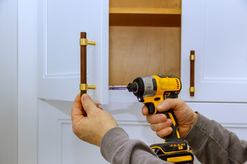 Worker uses power drill to attach cabinet handles in stylish kitchen, during modern kitchen...