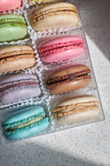 Bright French Macarons with white paperpan, plastic transparent packaging box on kitchen countertop, colorful cakes various flavors. appetizing dessert macaroons. Top down view