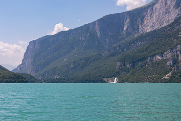 Small waterfall at alpine lake Lago di Molveno surrounded by steep cliffs and mountain peaks of majestic mountains Brenta Dolomites, National Park Adamello Brenta, Trentino, Italy. Lush green hills