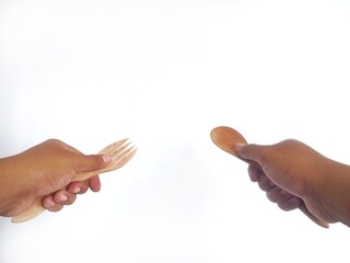 two hands holding wooden utensils: one hand holds a wooden fork with three prongs, while the other hand holds a wooden spoon. The utensils showcase a natural wood finish, emphasizing their handcrafted