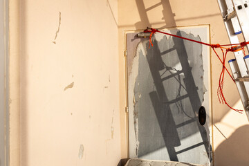 The angular shadow of a collapsible steel ladder stretches across a yellow wall, creating sharp...