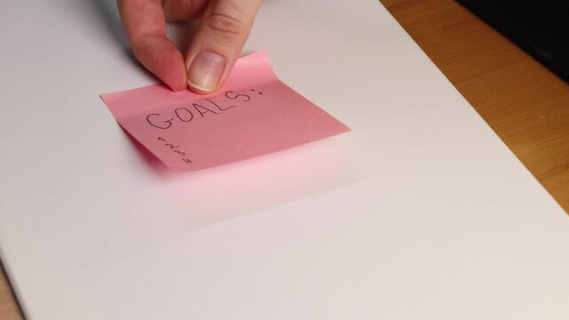 Hand sticking pink sticky note with word &ldquo;GOALS&rdquo; written on it. Symbol of planning and motivation. Close-up highlights goal-setting concept and future aspirations.