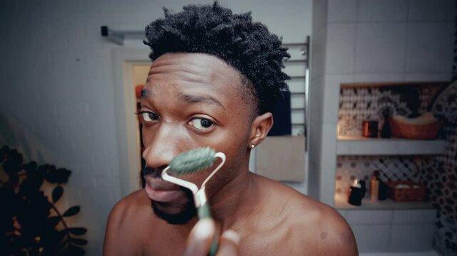 Young Black man standing shirtless in bathroom, looking into camera as massaging his face with jade roller during skincare routine. Close-up shot