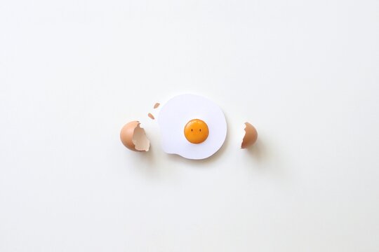 Overhead view of a conceptual fried egg with eyes and a cracked eggshell on a white background