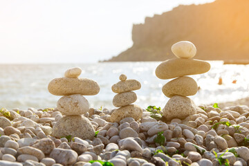 Three balanced stone stacks on a pebbly beach with a sea and rocky cliff in the background. The warm sunlight enhances the peaceful and serene atmosphere of this coastal scene