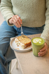 
A cosy café scene with a person in a green sweater holding a matcha latte. On the table, an almond-topped cake slice is served. Soft natural light. Minimalist style.