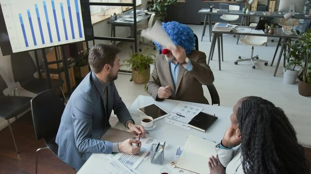 High angle view of hilarious boss in clown wig and nose berating and hitting laughing male colleague with documents while holding team meeting in office