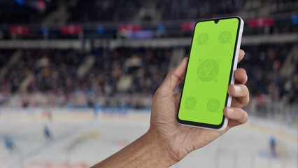 GREEN SCREEN CHROMA KEY A hand holding a phone with a green screen at a hockey game in an indoor stadium, ideal for showcasing live sports or betting apps