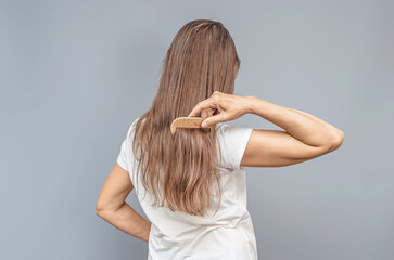 Fototapeta premium Wooden comb. Combing natural long hair. Rear view of woman in white t-shirt. Gray background, isolated.