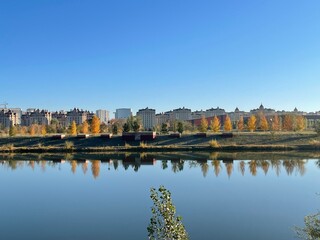 Calm river with a smooth surface in the foreground, concrete flowerbeds on the steep opposite shore, and rows of low-rise European-style buildings behind yellow autumn trees