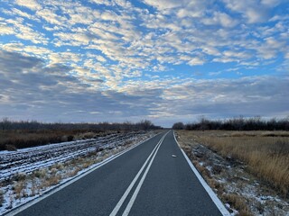 Early winter landscape with a gray asphalt road marked with white lines stretching into the horizon, flanked by a plowed field covered in first snow, leafless bushes, dry grass, and a cloud-filled blu