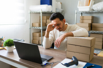 Stressed latin entrepreneur managing online orders with laptop and boxes, struggling with ecommerce...