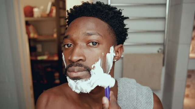 Young Black man with towel over his shoulder, looking into camera and using razor to shave face in bathroom. Close-up view
