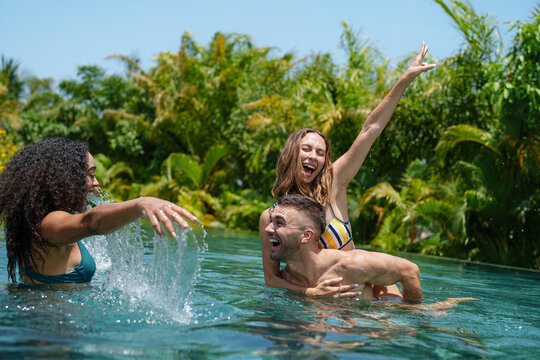 A diverse trio of cheerful young friends engage in playful splashing, expressing delight in a sunny pool setting, clad in swimwear.