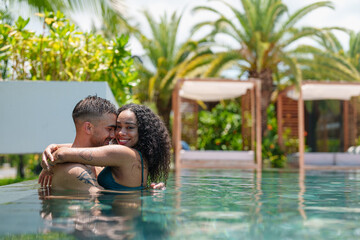 A young diverse couple basks in delight, exchanging loving gazes while embracing in a serene pool, with tropical palms enhancing the tranquil setting.