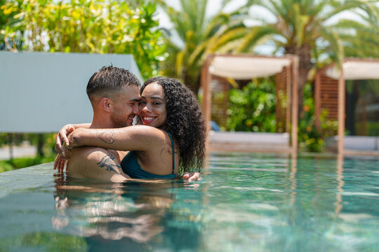 A diverse young adult couple enjoys an affectionate moment, with the woman's arms around the man, sharing a loving gaze in a sunlit swimming pool.
